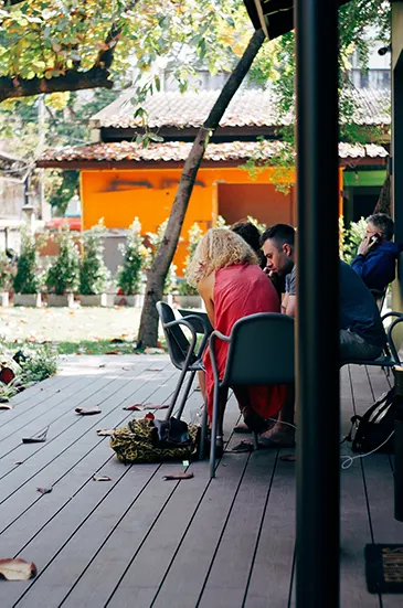 Drei Menschen arbeiten an Laptops auf einer schattigen Holzterrasse unter Bäumen