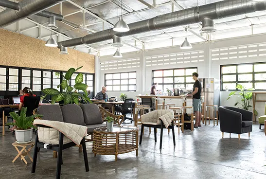 Loungebereich im Coworking mit Korbmöbeln, Pflanzen und Lüftungsrohren unter der Hallendecke