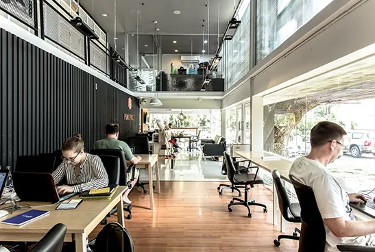 Coworking-Raum mit Holztischen, mehreren Arbeitenden und Galerie über der Fensterfront