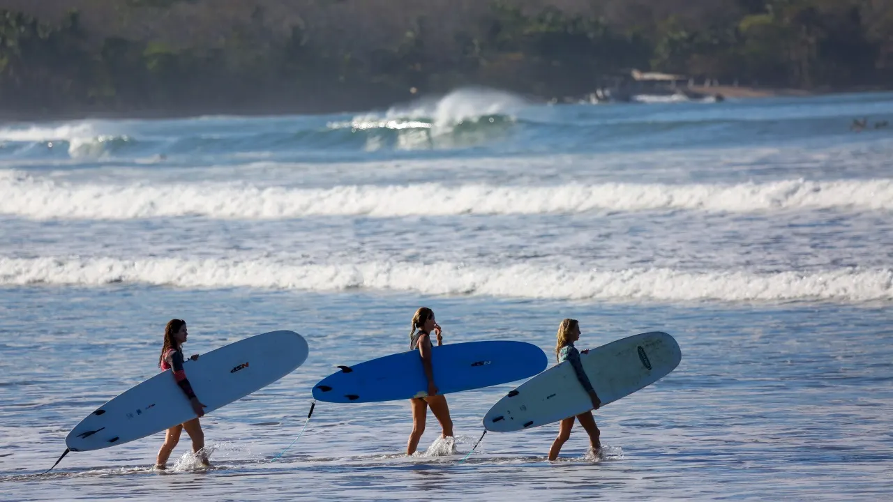 Drei Surferinnen laufen mit Longboards durchs flache Wasser, Brandung im Rücken