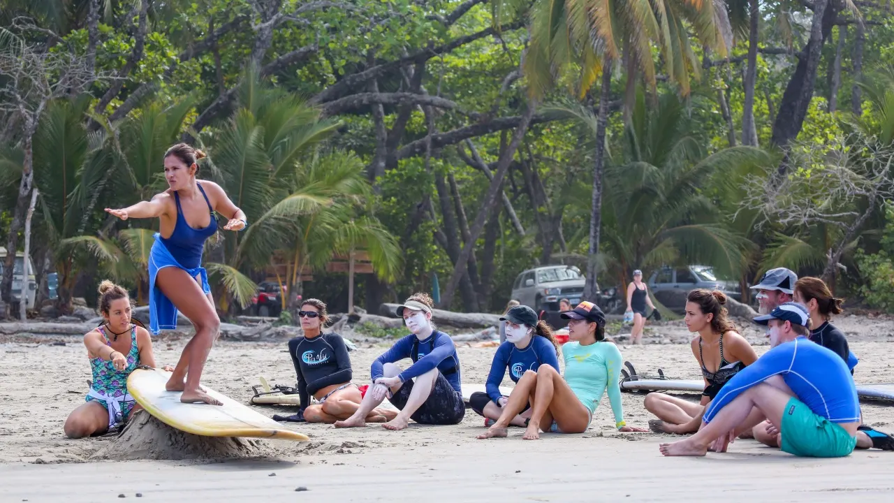 Surflehrerin zeigt auf einem Board am Strand die Aufstehbewegung, die Gruppe sitzt im Sand