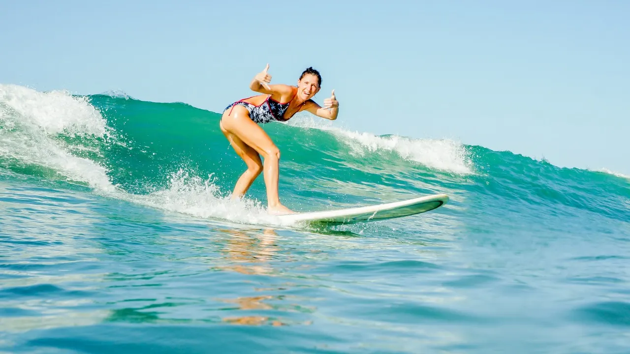 Surferin zeigt beide Daumen nach oben, während sie eine türkise Welle abreitet