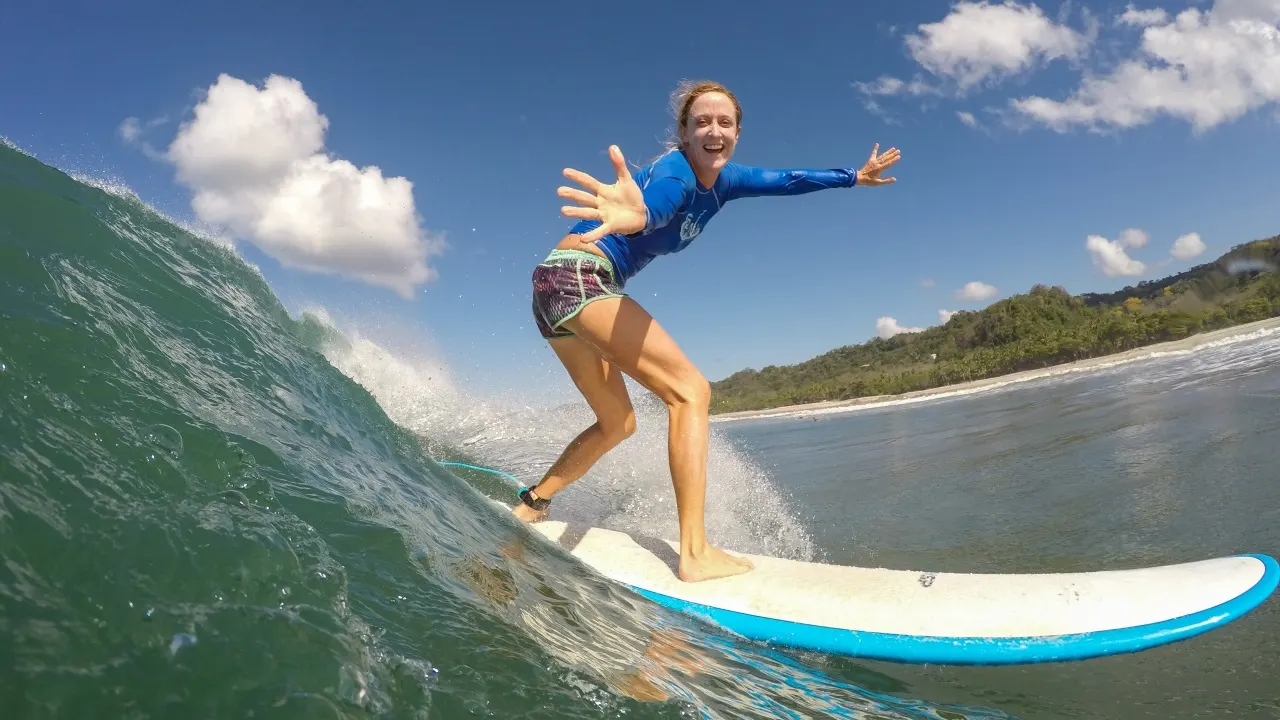 Lachende Surferin in blauem Longsleeve balanciert mit ausgebreiteten Armen auf dem Board