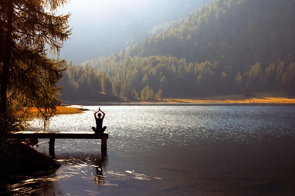 Silhouette einer meditierenden Person auf einem Holzsteg am Bergsee im Gegenlicht