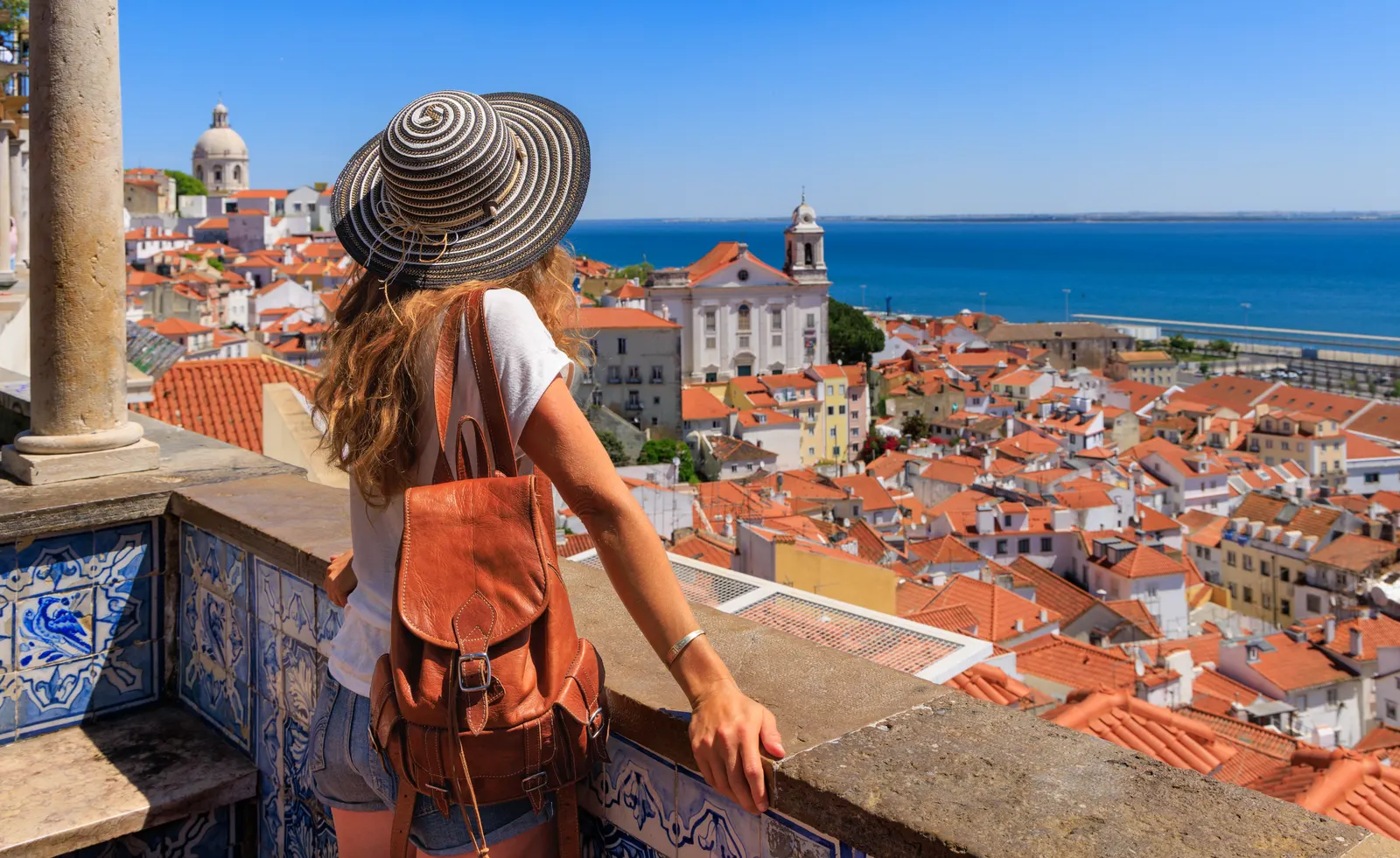 Reisende mit Strohhut und Rucksack blickt von einer Aussichtsterrasse auf eine mediterrane Altstadt mit roten Dächern am Meer