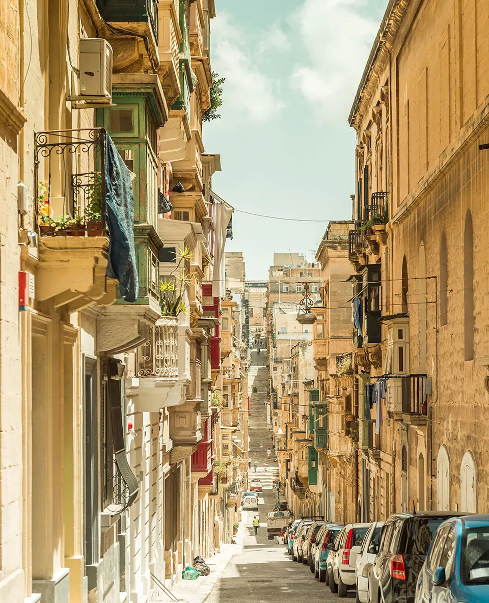 Steil abfallende Gasse mit vorspringenden Balkonen, Wäsche und parkenden Autos in Valletta