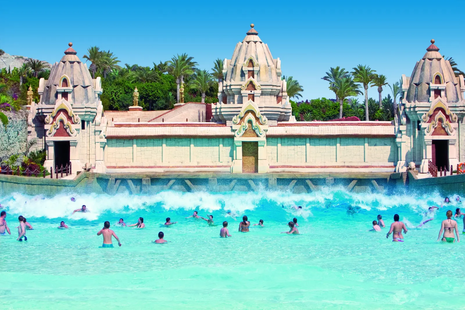 Brechende Welle im Wellenbecken des Siam Park vor thailändischer Tempelfassade mit vielen Badenden