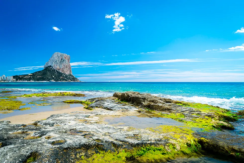 Algenbewachsene Felsen am flachen Ufer, dahinter türkises Meer und der Peñón de Ifach