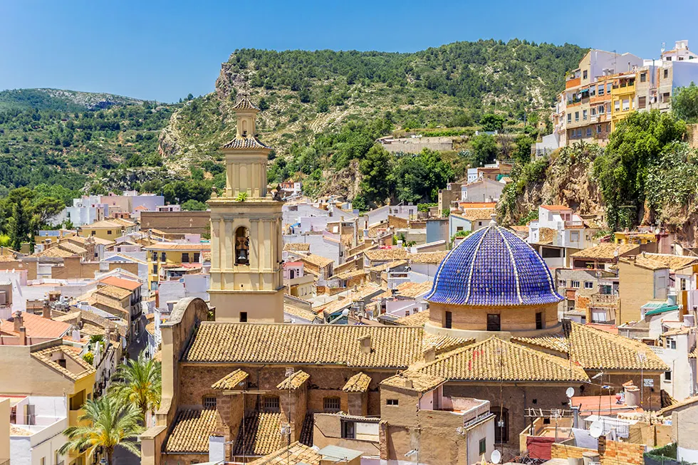 Ortsansicht von Buñol mit Kirchturm, blauer Kuppel und Ziegeldächern vor bewaldeten Hügeln