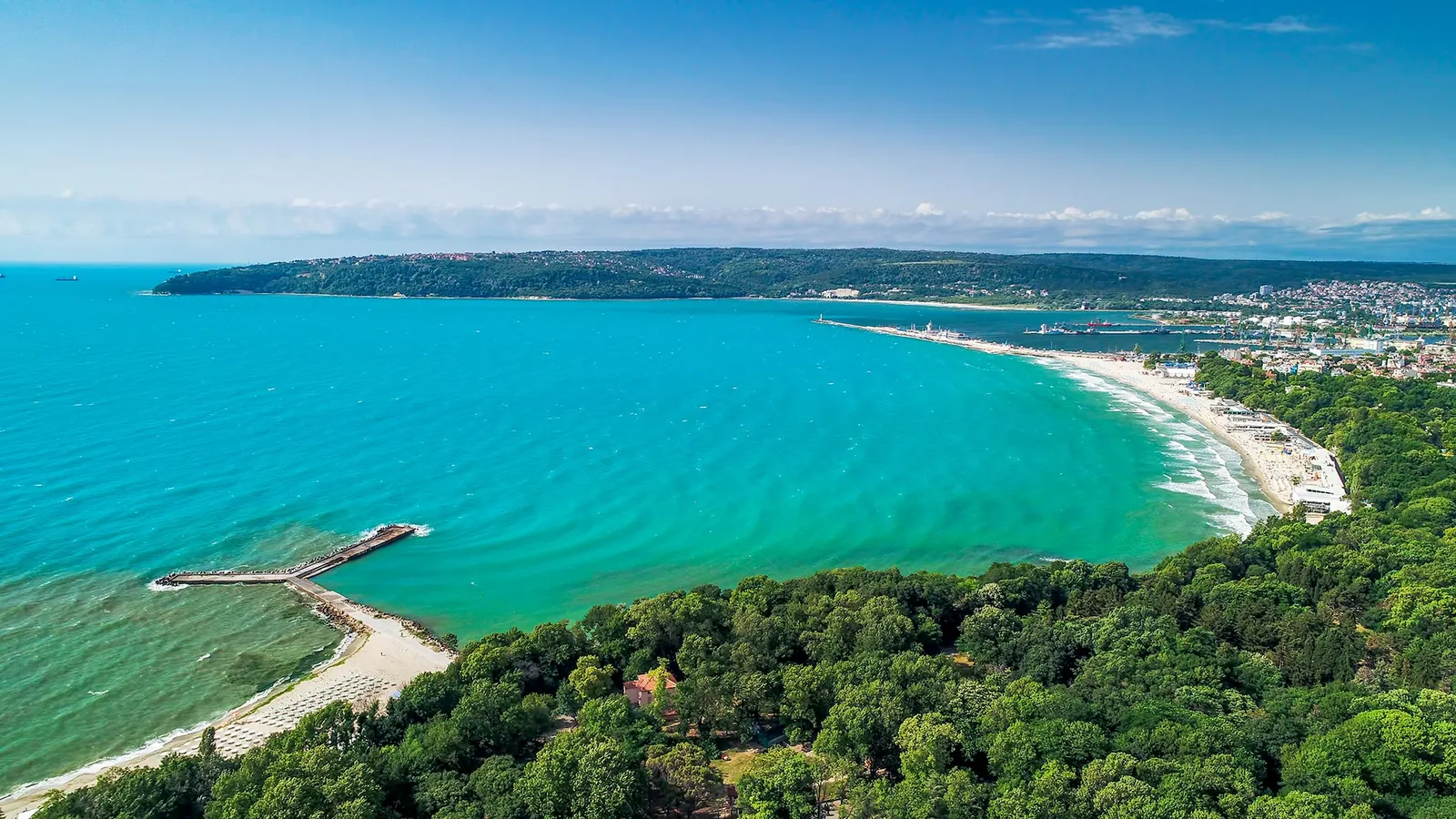 Luftaufnahme einer Bucht mit türkisem Meer, bewaldetem Ufer, Sandstrand und Seebrücke
