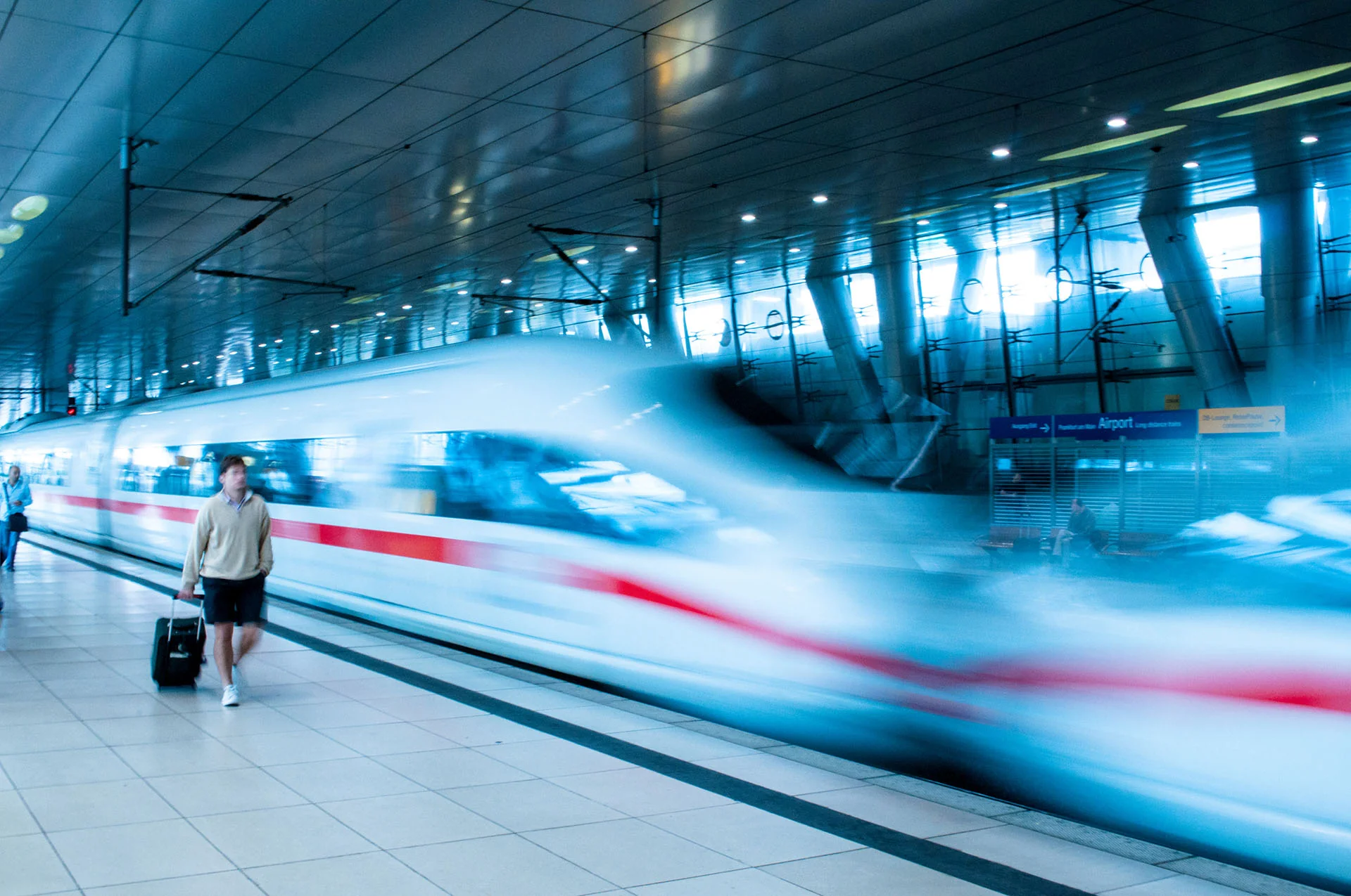 ICE-Zug fährt mit Bewegungsunschärfe am Fernbahnhof Frankfurt Flughafen ein, ein Reisender mit Koffer wartet auf dem Bahnsteig