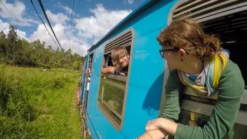 Zwei Reisende lehnen sich aus der offenen Tür eines blauen Zuges in grüner Landschaft
